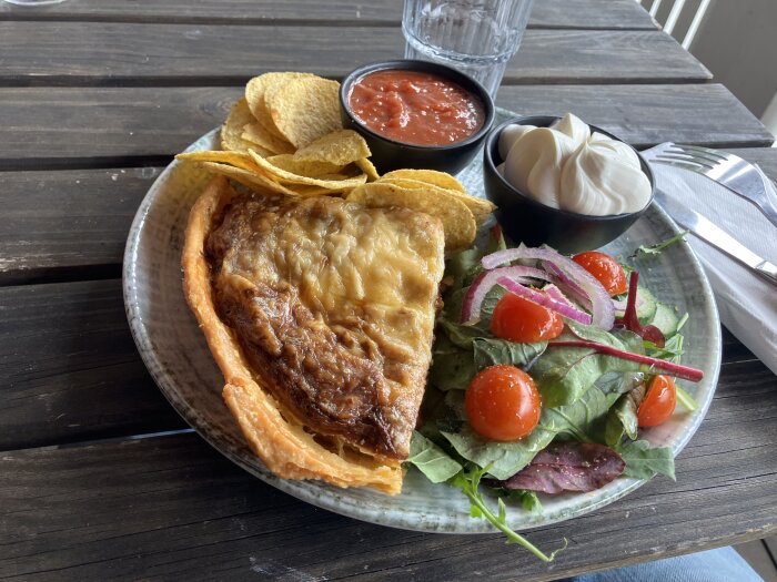 Tallrik med quiche, nachos, salsa, crème fraiche och sallad med körsbärstomater och rödlök på ett mörkt träbord.