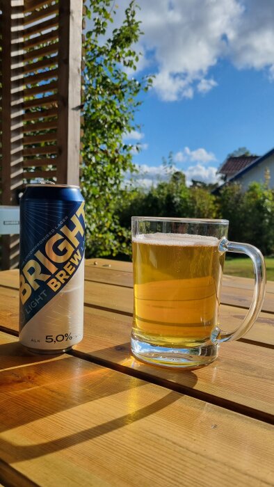 En burk och ett glas ljus öl står på ett träbord utomhus en solig dag.