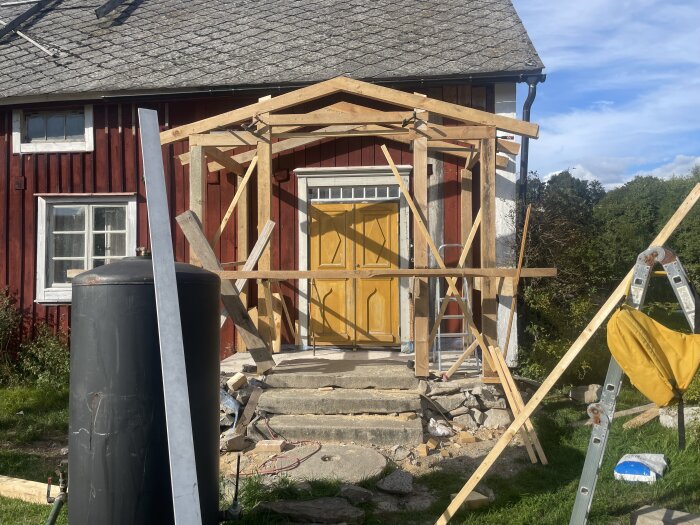 Byggen av veranda på rött hus med gult dörr, takstolar, och diverse verktyg runtom.