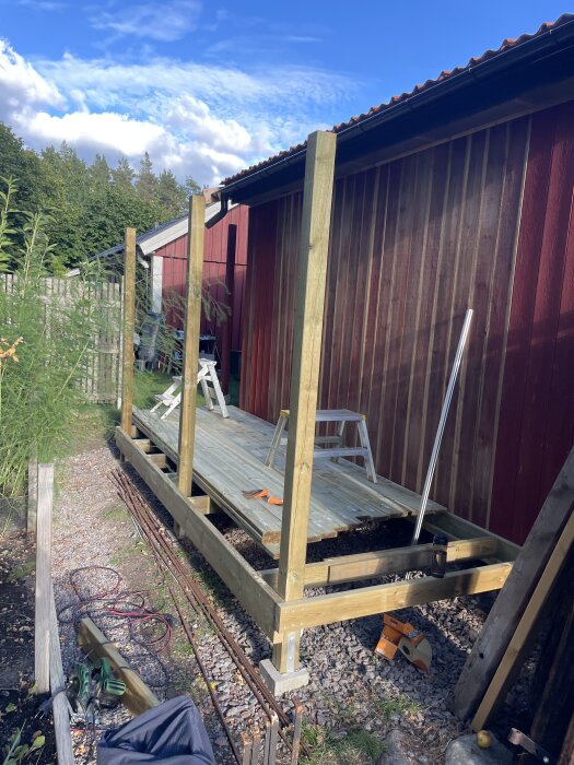 Träbjälklag under konstruktion för en trädgårdsbod, med golvbalkar av spillvirke bredvid röd byggnad och stege i bakgrunden.