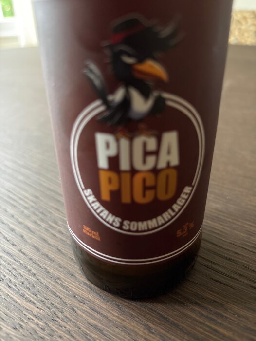 Brun flaska med etikett för "Pica Pico Skatans Sommarlager", 5,3% alkoholhalt, ser ut som en vanlig läskflaska på träbord.