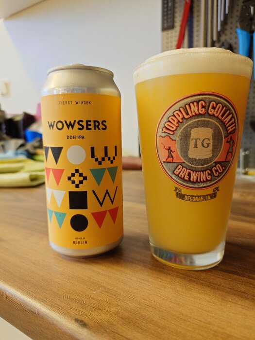 En burk Wowsers DDH IPA och ett glas med öl från Toppling Goliath Brewing Co. står på ett träbord.