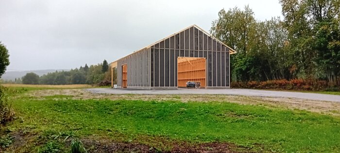 En nybyggd hall med trästomme på en grön tomt kantad av träd och växtlighet.