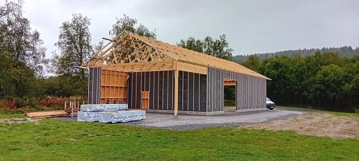 Halvbyggt trähus med bjälkar och väggpaneler, omgiven av skog och gräsmatta, med material upplagda bredvid.