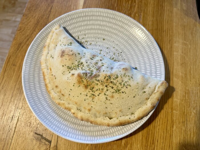 En calzone på en tallrik med örtkryddor ovanpå, fotad på ett träbord.
