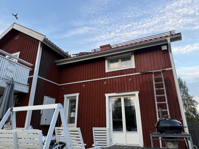 Rött trähus under målning, vit veranda, stege mot väggen, grill och utemöbler i förgrunden, blå himmel med moln.