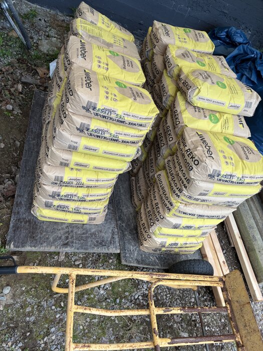 Staplar av Weber grovbetong i gula säckar på lastpallar utomhus, med en gul rostig skottkärra i förgrunden.