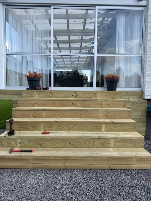 Trätrappa med infällda decklights, omgiven av grus; verktyg och två krukor med växter på trappan, framför glasad vägg.