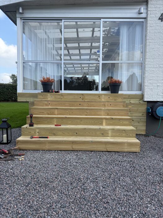 Trappa byggd av trätrallbrädor med infällda decklights, kompletterad med verktyg och växter, framför en inglasad uteplats.