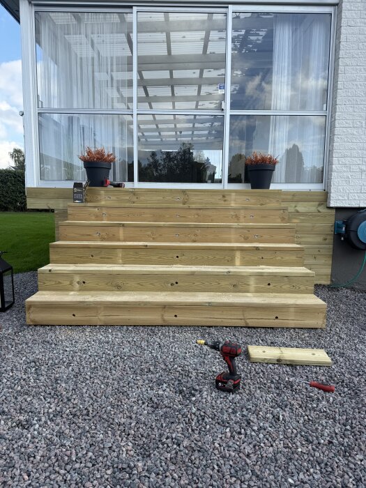 Trätrappa med 18 infällda decklights, omgiven av stenläggning, framför en glasad altan. En borrmaskin och träplanka syns på marken.