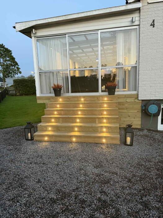 Trätrappa med infällda decklights framför inglasad altan, omgiven av grus och dekorerad med växter och lyktor.