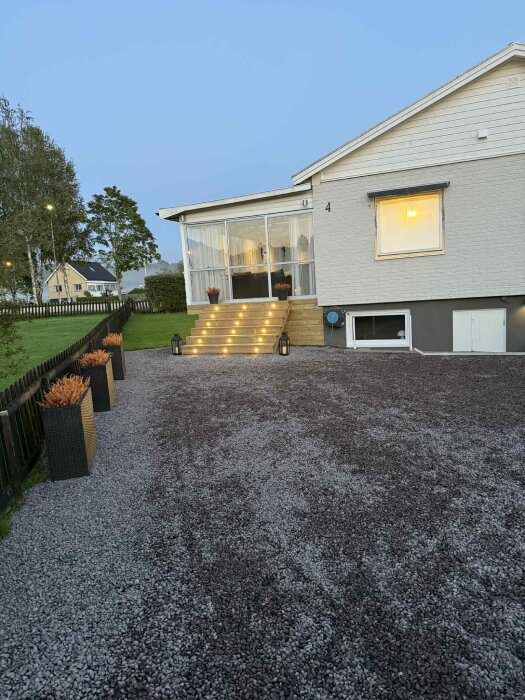 Trappa med infällda decklights leder upp till entré, omgiven av krukor och grusad gångväg, skapande en kvällseffekt på en altan.