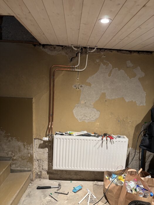 Vit radiator monterad på en rå källarvägg med synliga koppar- och plasrör ovan. Verktyg ligger ovanpå och runt radiatorn, inklusive hammare och skiftnyckel.