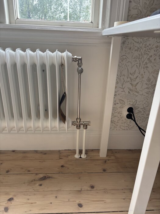 Vit radiator installerad vid ett fönster med rör dragna längs väggen och golvet, del av en renovering med fokus på golvvärme och radiatorer.