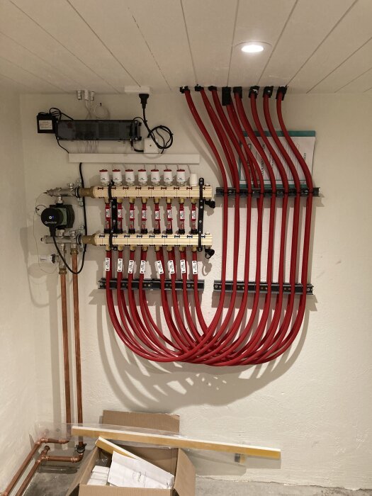 Golvvärmeinstallation i källare, visar kopplingspanel med flera röda rör och kopplingar, samt en pump på väggen.