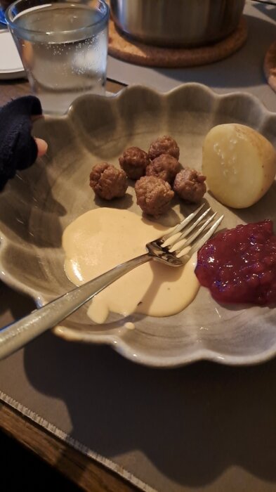 Köttbullar, gräddsås, lingonsylt och potatis på tallrik med glas vatten vid sidan.
