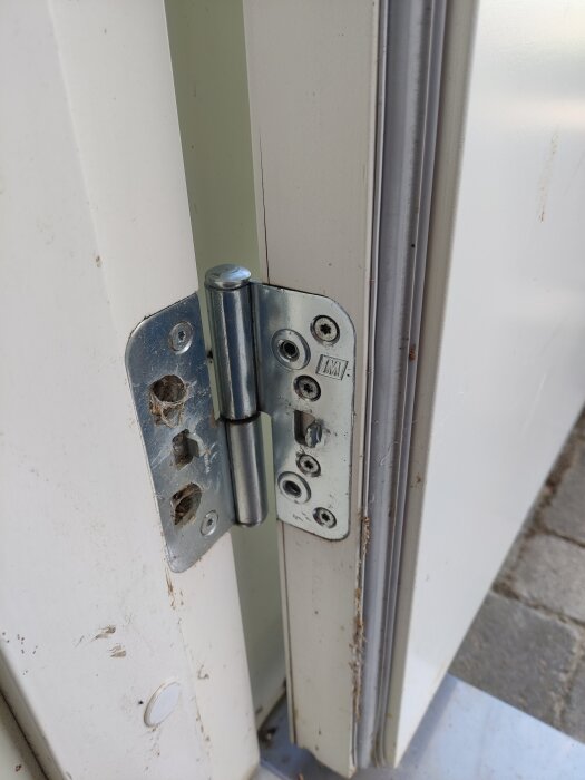 Ytterdörrens gångjärn med utbytt skruv som möjliggör justering; synlig glipa vid dörrbladets nederkant.