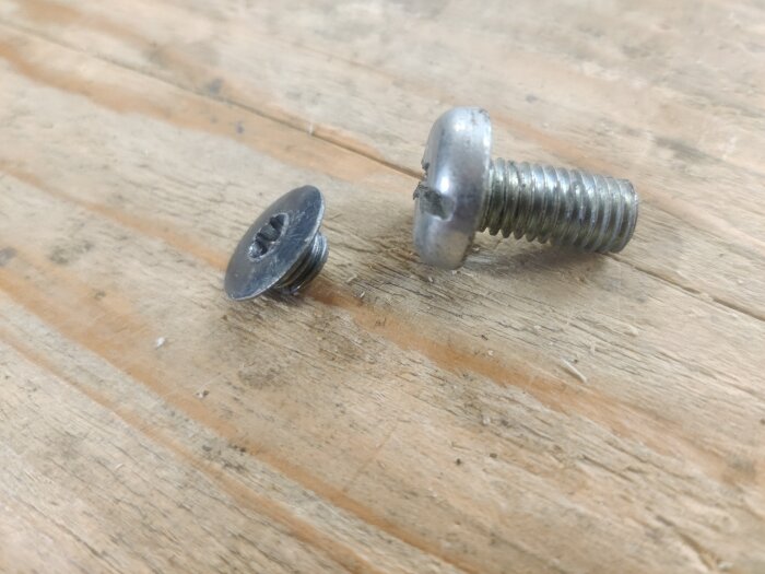 Två skruvar ligger på en träyta. Den ena är silverfärgad med spårskruvhuvud, den andra är mörkare med insexformad topp.