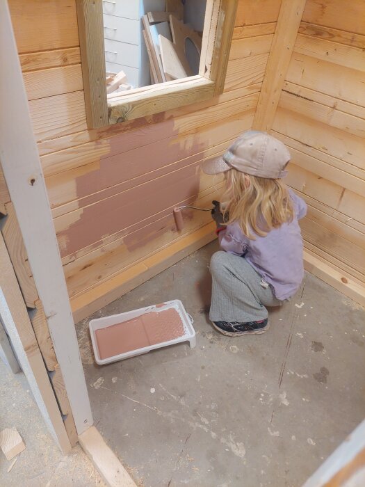 En liten person med keps målar en vägg i ett träskjul med roller och rosa färg. Målarbytta med färg står på golvet.