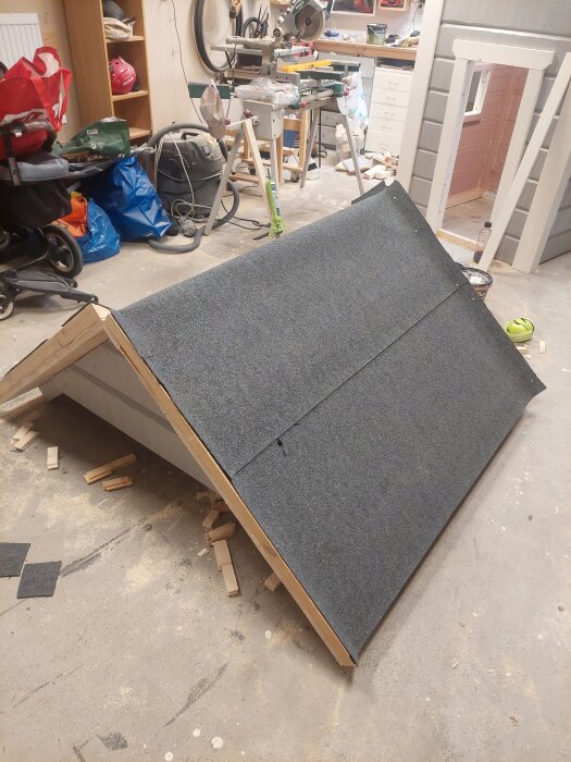 Litet modellhus under byggnation i garage, taket har svart takpapp och golvet är stökigt med träbitar och verktyg runt omkring.