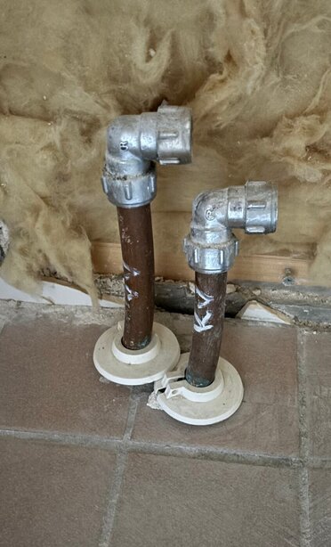 Två metallrör med böjda kopplingar sticker upp från golvet framför en vägg med isolering synlig.