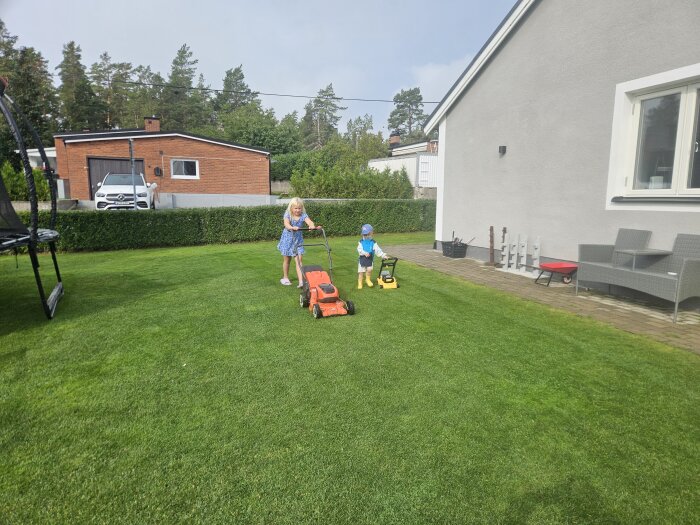 Två barn klipper gräsmattan framför ett hus med varsina gräsklippare, en större orange och en mindre gul, med en studsmatta i bakgrunden.