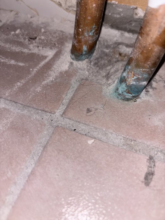 Kopparrör i ett badrum med ansamling av grönt oxidationsrester och damm vid skarven på kaklat golv.