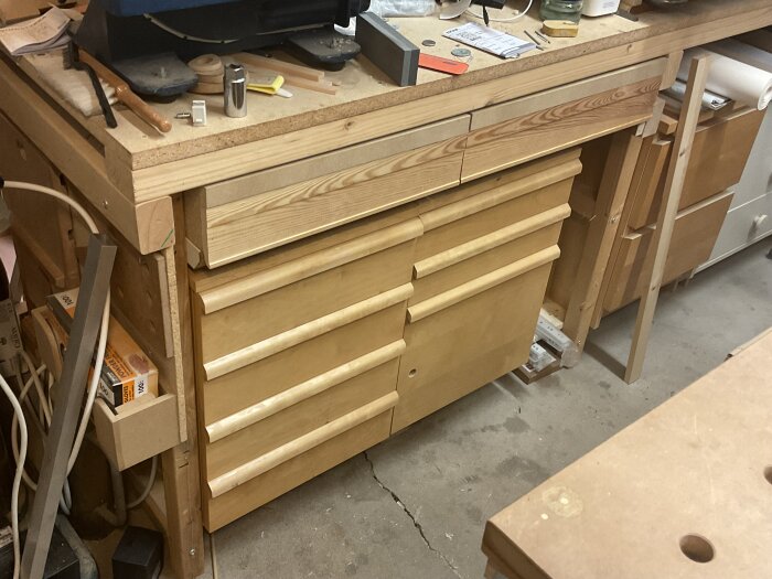 Gedigen verkstadsbänk med trälådor och verktyg ovanpå, hyllor undertill, omgiven av diverse arbetsmaterial.