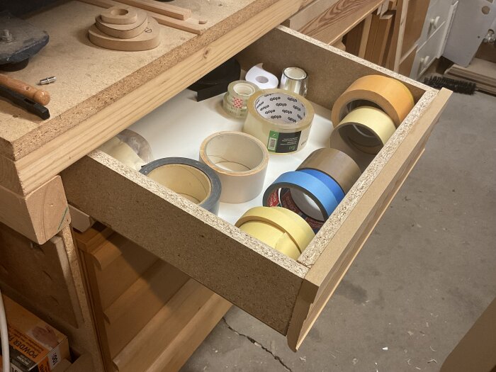 Öppen låda av spånskiva med olika sorters tejprullar inuti, synlig del av arbetsbänk i trä ovanför.