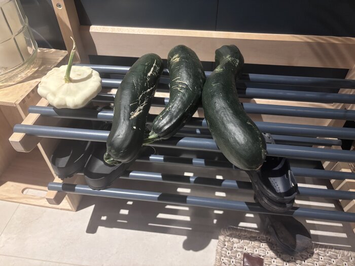 Tre zucchinis och en vit pumpa ligger på ett skoställ i trä med ljusgråa metallribbor.
