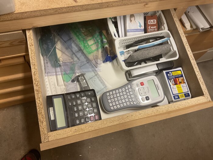 Öppen låda med olika kontorsmaterial, inklusive en miniräknare, en etikettskrivare, pennor, linjaler och tejpkassetter.