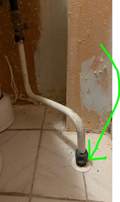 Närbild på röranslutning i ett badrum, markerad med en grön pil, mot en beige vägg med synliga kakelplattor.