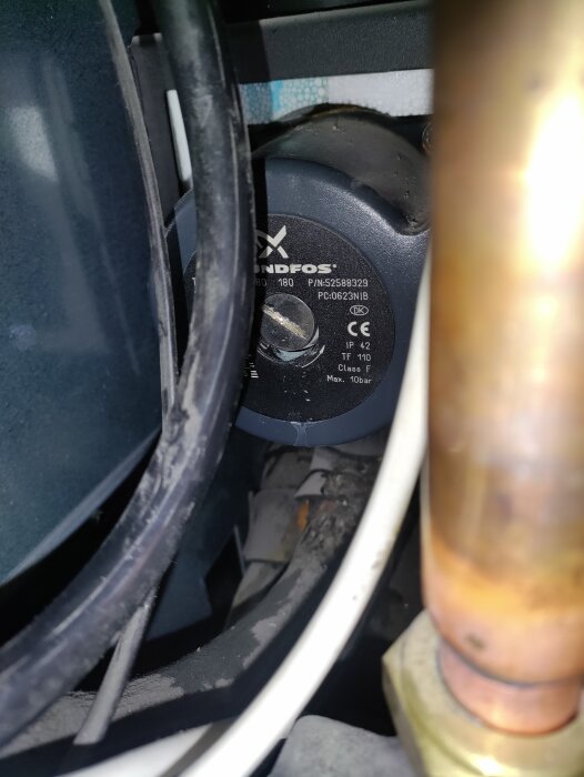 Pump med märkning "Grundfos" och komponenter inuti ett rörsystem, delvis skymd av kablar och rör, möjlig justeringspunkt med skruvmejsel.