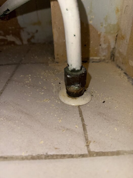 Vitt rör som går genom ett kaklat golv, omgiven av smuts och damm, med en metallisk koppling vid basen.