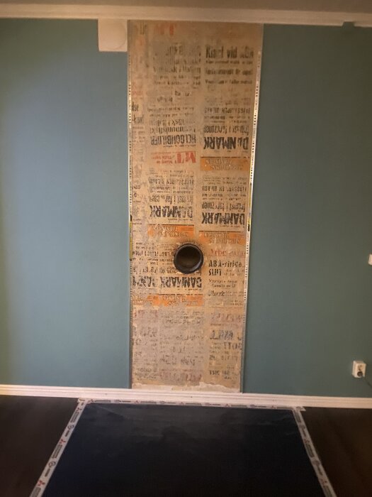 Gammaldags tapet med text synlig på väggen efter att modern tapet tagits bort, omgiven av målade väggar, med ett rör som genomborrar tapeten.