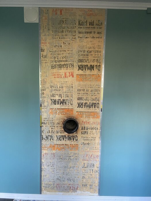 Vägg med gammalt tidningspapper och hål för skorsten, upptäckt bakom tapet, omgiven av blå väggar.