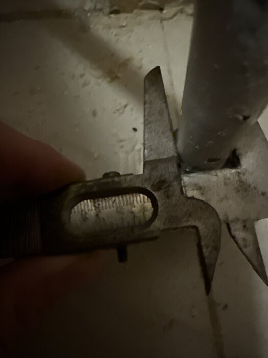 A caliper measuring the diameter of a metal pipe against a tiled background.