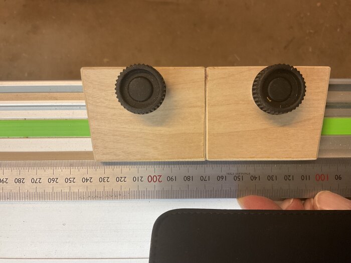 Trästycken med svarta knoppar monterade på skena med grön linje och linjal som visar centimeter och millimeter.