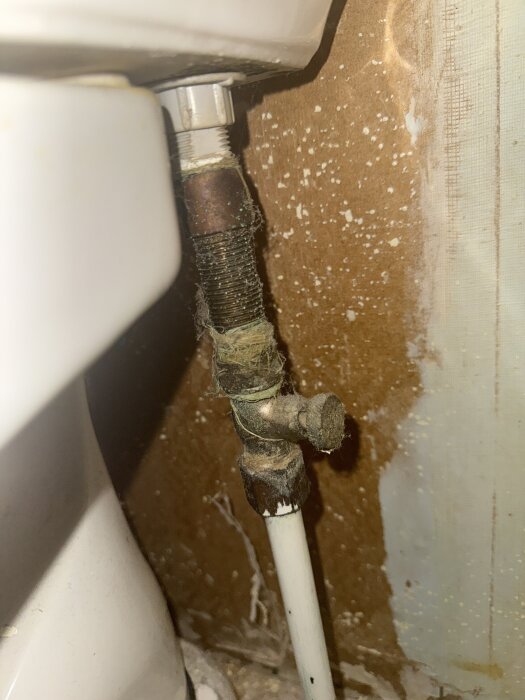 Rör och ventil nära en toalett monterad mot en brun vägg, ytorna är dammiga och slitna.