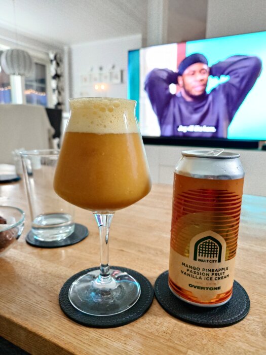 Ett glas gul suröl med skum och en ölburk på ett bord, i bakgrunden syns en tv-skärm.