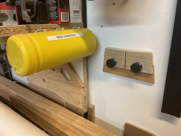 Gul behållare märkt "Ren handduk" monterad på väggen i verkstad, intill träpanel med två svarta knoppar.