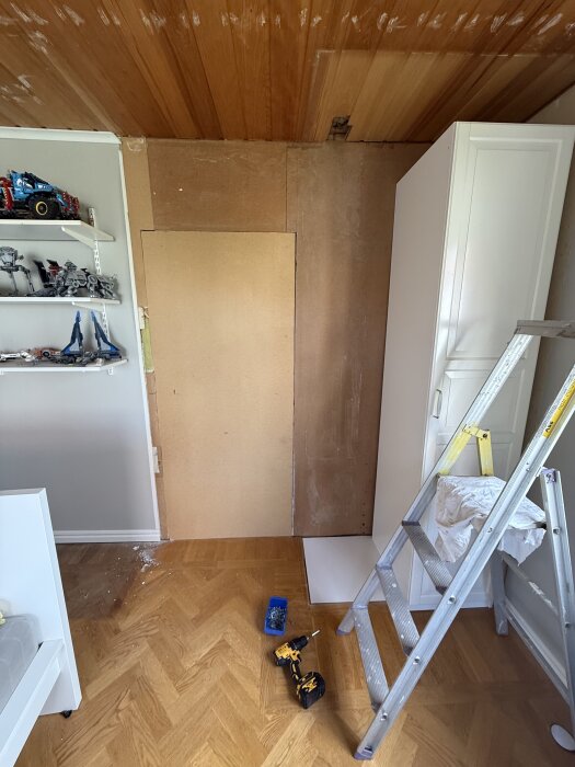 Barnrum under renovering med exponerad masonitvägg, byggplyfa och ny garderob. Stege, borrmaskin och leksakshyllor syns i rummet.
