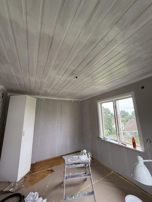 Rum under renovering med nyligen målat tak, stege, och plastskyddat golv, samt ett fönster med utsikt över grannskapet.