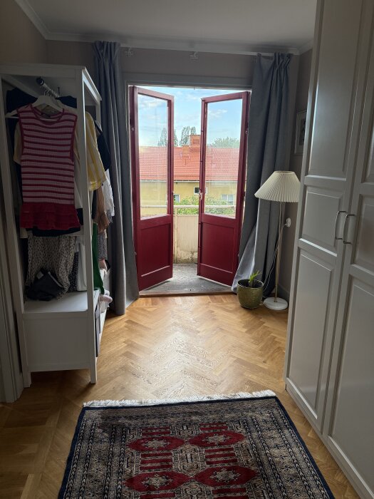 Ett sovrum med parkettgolv i fiskbensmönster, garderober, en matta, och öppna balkongdörrar med utsikt mot ett rött tak.