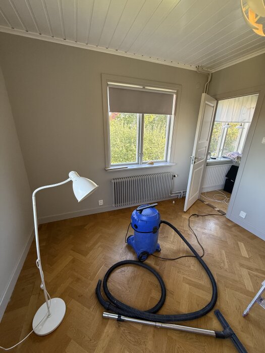Ett renoverat rum med parkettgolv, en blå dammsugare, vit golvlampa och öppna balkongdörrar. Hålrummet i golvet visar pågående arbete.