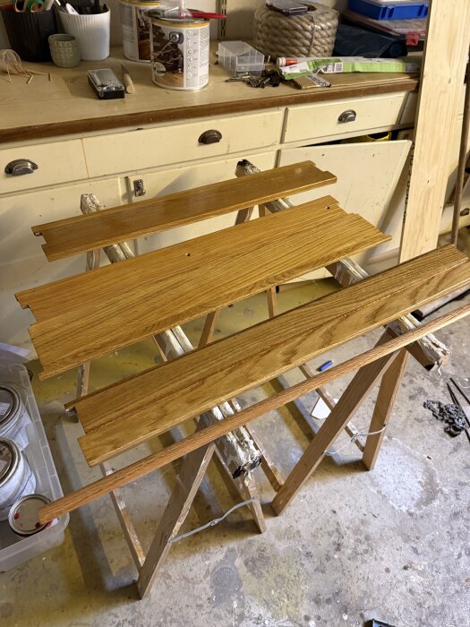 Lackerade träplankor på bockar i ett verkstadsutrymme med verktyg och målarutrustning på bänken i bakgrunden.