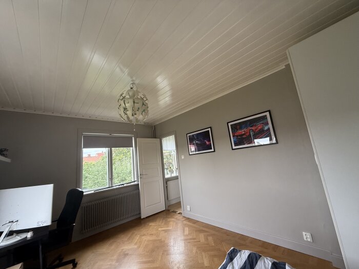 Nyrenoverat rum med vitmålat tak, ljusgrå väggar, två tavlor, skrivbord och svart stol. Trägolv i fiskbensmönster och utsikt genom fönster.