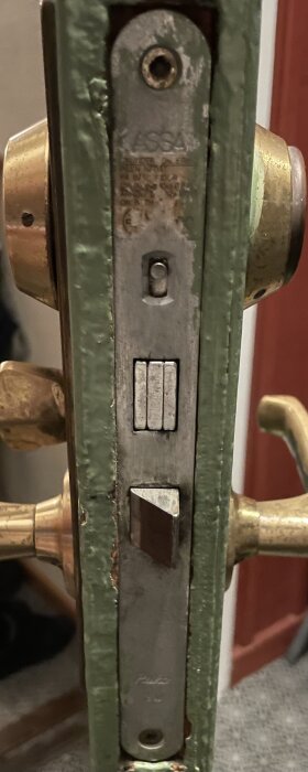 Närbild av en grön dörrkarm med ett monterat dörrlås av märket Assa. Låset har synliga detaljer och patina.