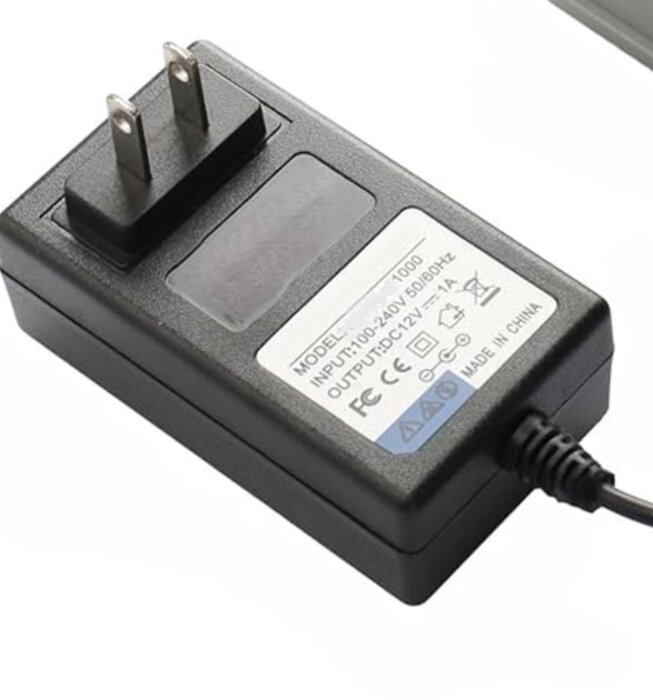 En svart strömadapter med 100-240V ingång; visar etikett med tekniska specifikationer och CE-märkning.