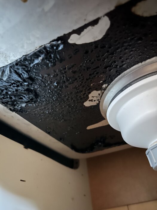 Svart ljuddämpningsmatta under diskho med stel, delvis lossnad yta och kvarvarande svart klister. En vit rörkoppling syns till höger.